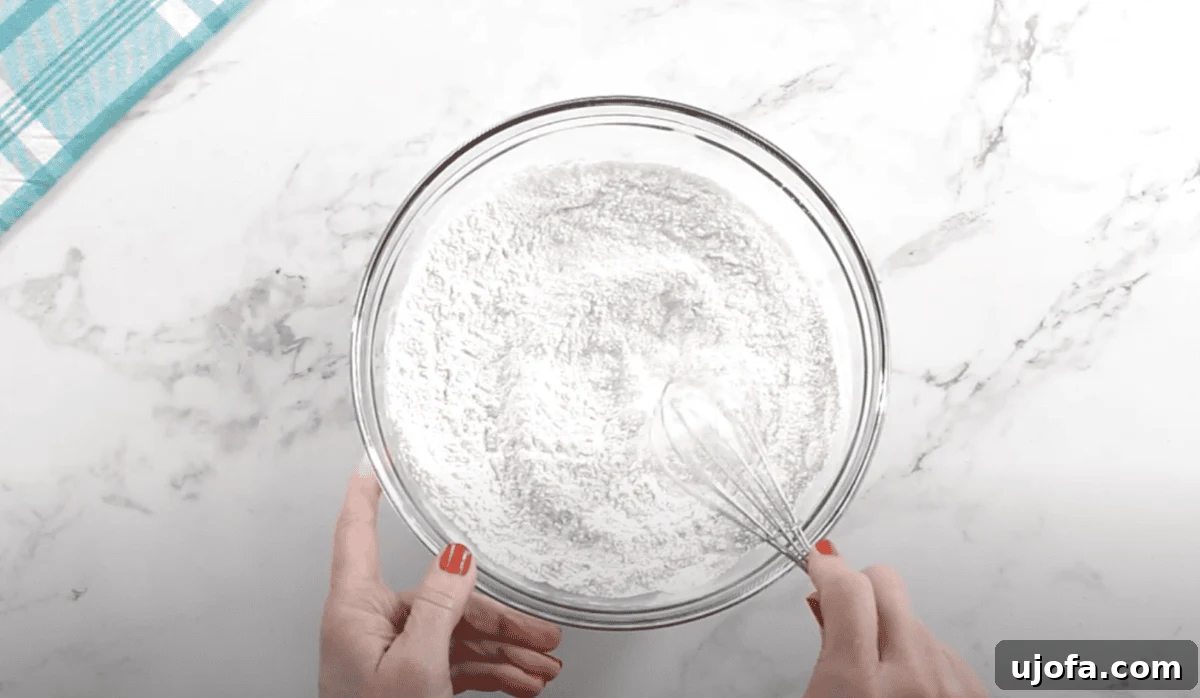Overhead view of a person whisking a mixture of dry ingredients—gluten-free flour, sugar, and yeast—in a large glass mixing bowl on a white marble surface.