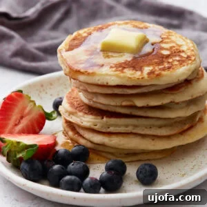 Gluten Free Brunch Delights 4 A stack of fluffy gluten-free pancakes topped with melting butter and drizzled with syrup, served on a speckled white plate with fresh strawberries and blueberries on the side. A dark napkin is in the background.