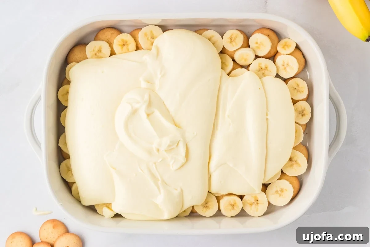 Half of the creamy banana pudding mixture spread over a layer of cookies and bananas in a white baking dish, ready to be smoothed out.
