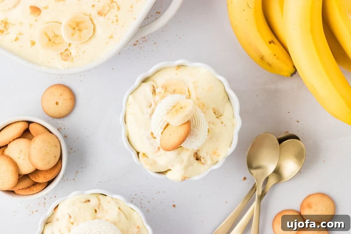 Flat lay of individual servings of gluten-free banana pudding in white bowls, garnished with whipped cream, banana slices, and cookies. Surrounded by extra cookies, fresh bananas, and gold spoons on a light background.