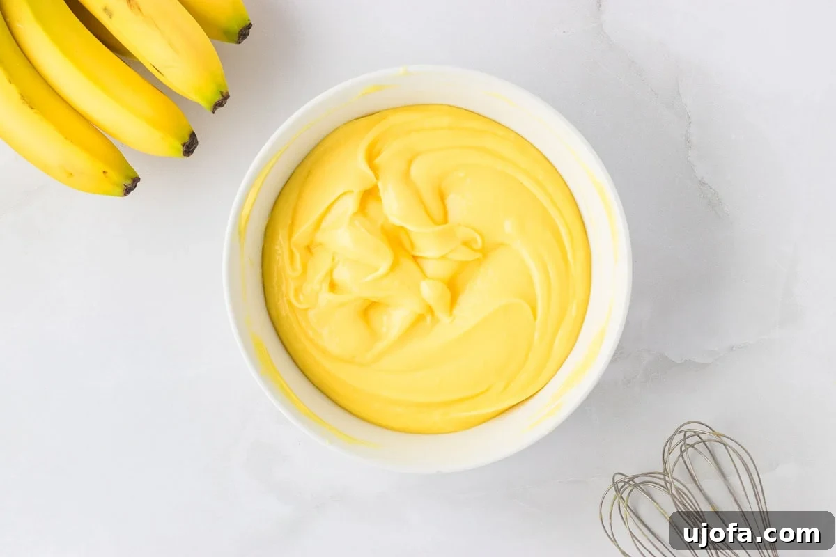 A white bowl filled with smooth, bright yellow vanilla pudding, with a bunch of bananas nearby on a light countertop.