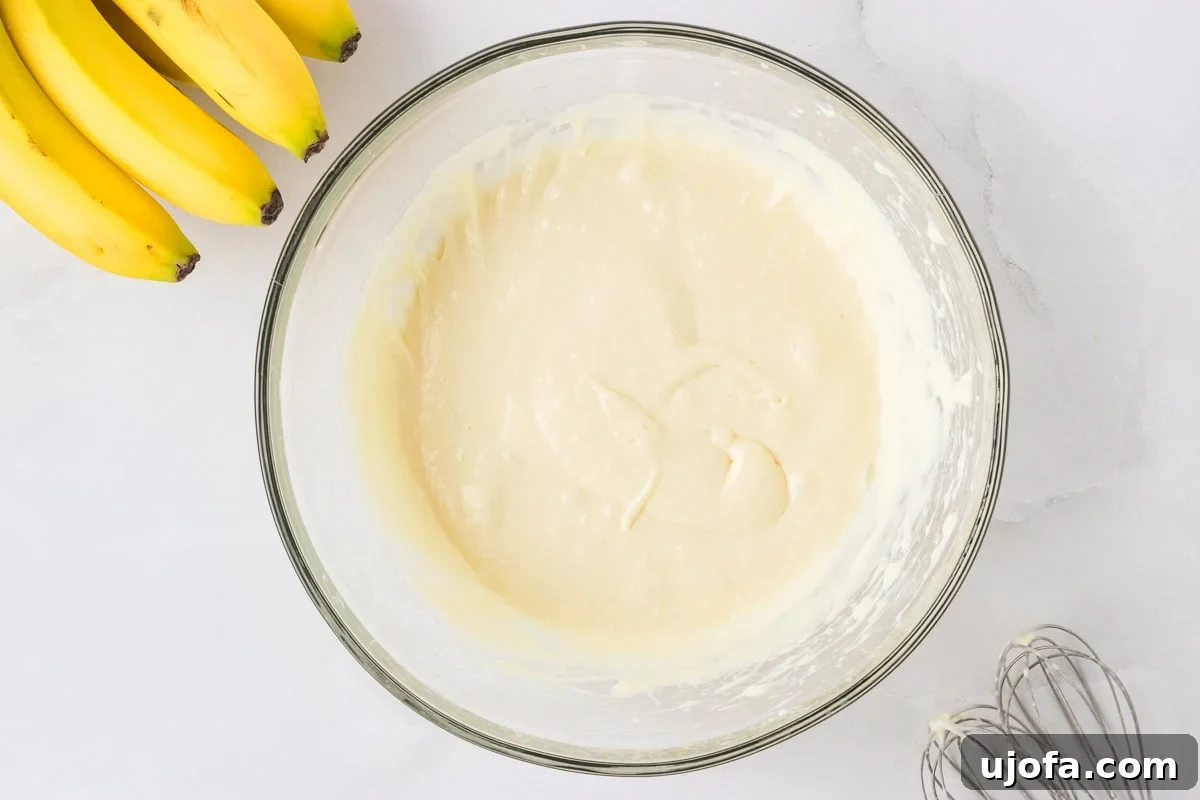 Creamy mixture of softened cream cheese and sweetened condensed milk in a glass mixing bowl, with bananas and a whisk in the background.