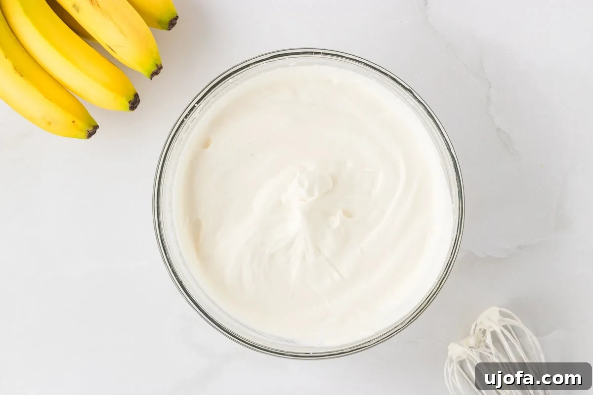 A bowl of freshly whipped cream with stiff peaks in a glass bowl on a white surface, bananas and a whisk nearby.