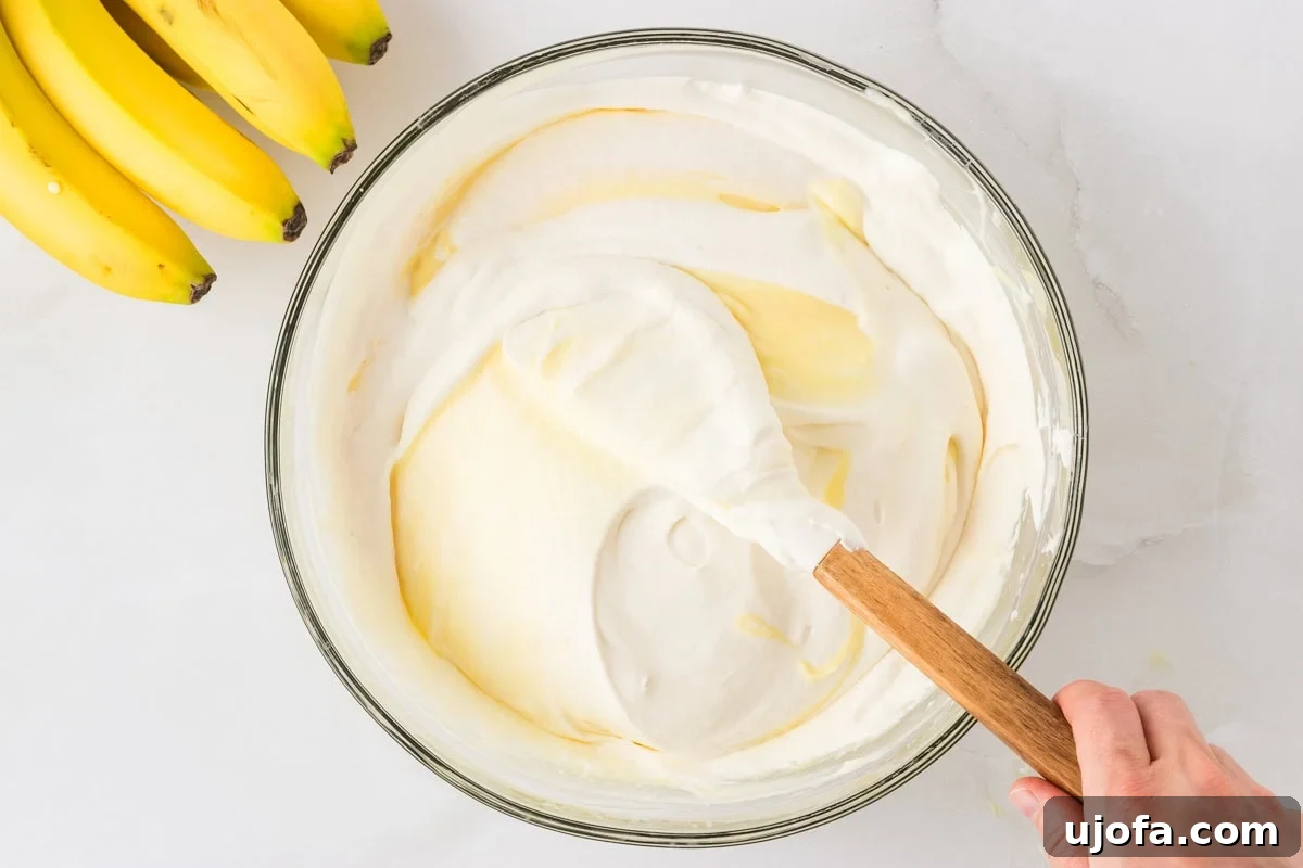 Pudding and whipped cream mixture being gently folded together in a glass bowl with a rubber spatula. Bananas are visible on the side.