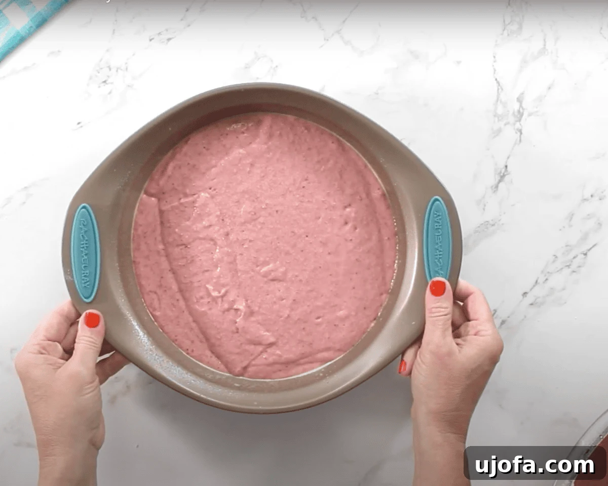 Luscious Strawberry Gluten-Free Cake 14 A round cake pan filled with smooth, pink strawberry cake batter. A person with red nail polish holds the pan steady on a white marble surface.