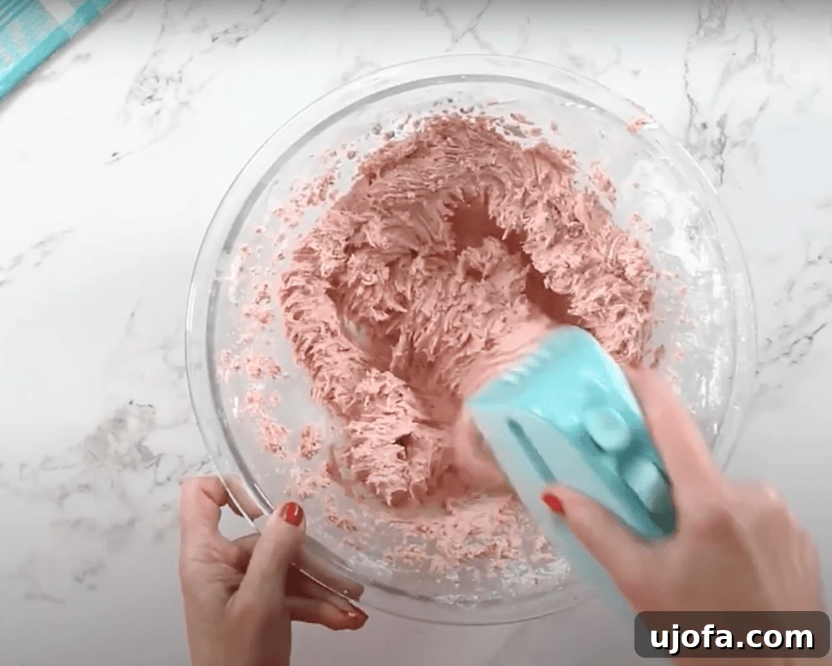 Luscious Strawberry Gluten-Free Cake 19 A glass mixing bowl with a pink strawberry buttercream frosting being mixed with a blue hand mixer. The frosting has a fluffy and creamy texture.