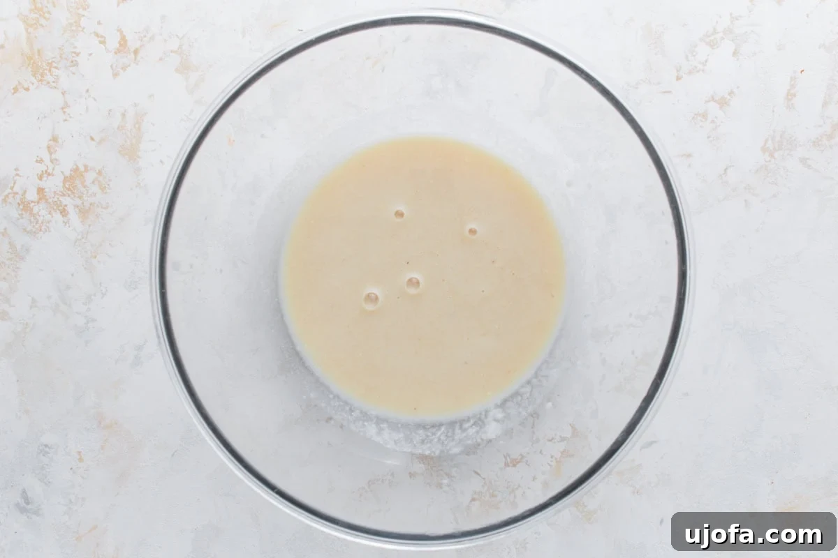 Clear mixing bowl with a smooth, beige liquid mixture of wet ingredients for oatmeal bars on a light textured background, including eggs, milk, applesauce, brown sugar, and vanilla extract.