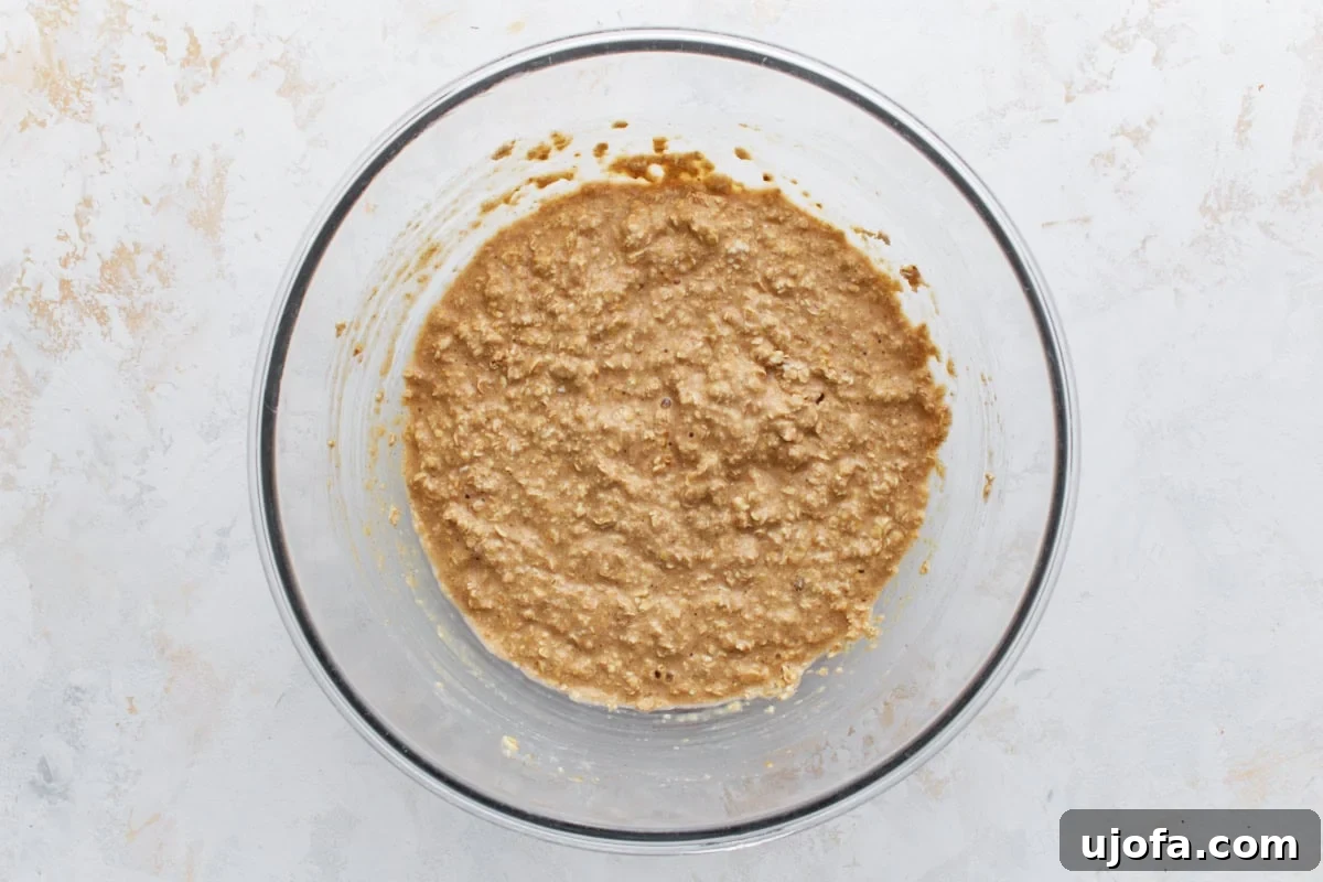 Wet and dry ingredients combined into a thick, textured oatmeal batter in a glass mixing bowl, showing the oats, baking powder, baking soda, salt, and cinnamon incorporated.