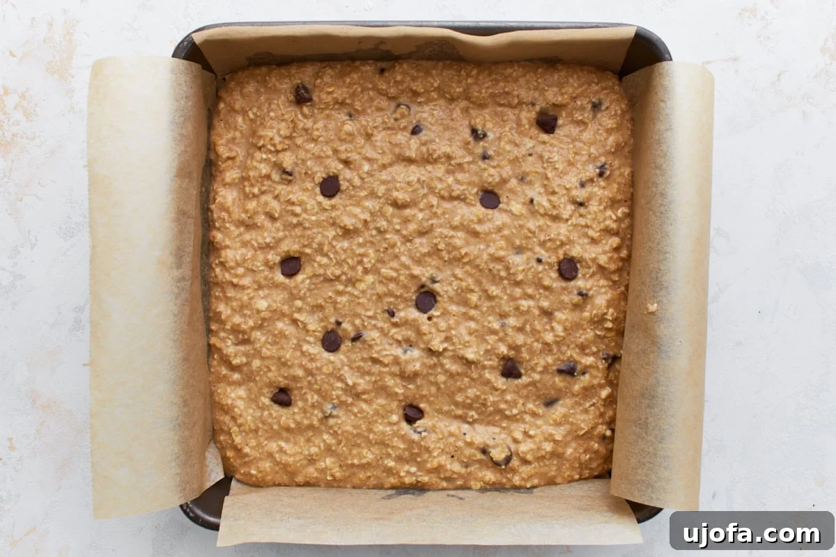Chocolate chip oatmeal batter spread evenly in a parchment-lined square baking pan before baking, showing a smooth top surface.