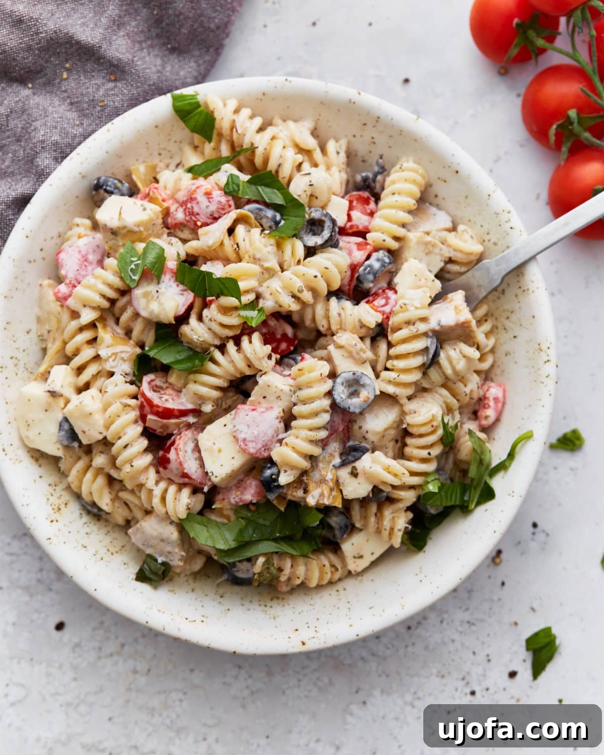 A vibrant bowl of creamy Italian pasta salad, artfully garnished with fresh basil leaves. The salad features perfectly cooked rotini pasta, halved cherry tomatoes, sliced black olives, delightful mozzarella cubes, and a rich, creamy dressing.