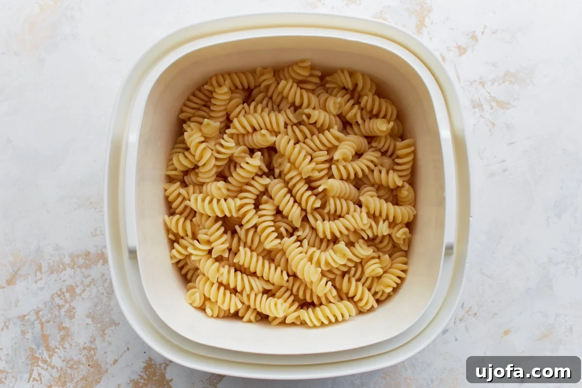 A close-up of plain cooked rotini pasta in a white plastic colander, showing its perfectly al dente texture, ready to be mixed with other ingredients.