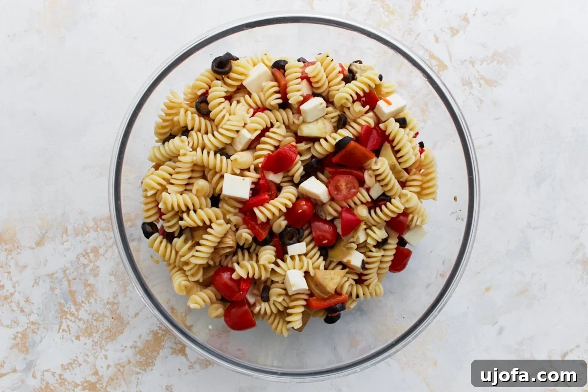 A large glass mixing bowl filled with a colorful array of cooked rotini pasta, chopped cherry tomatoes, black olives, vibrant roasted red peppers, tender artichoke hearts, and diced mozzarella cubes, waiting for the dressing.