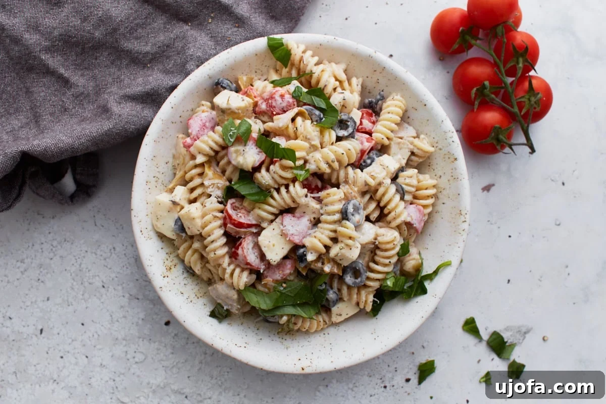 A top-down artistic view of a beautifully prepared bowl of creamy Italian pasta salad, strategically placed beside a cluster of fresh tomatoes on the vine and a rustic linen napkin, emphasizing its freshness and appeal.