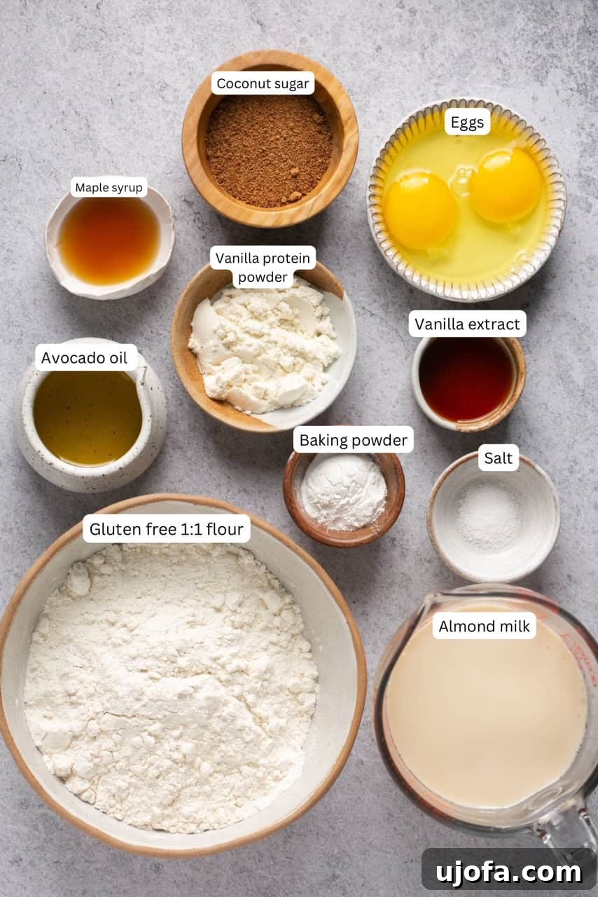 A flat lay of small bowls containing the labeled ingredients for gluten-free waffles: flour, baking powder, vanilla protein powder, almond milk, coconut sugar, maple syrup, vanilla extract, eggs, avocado oil, and salt.