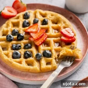 A gluten-free waffle on a pink plate topped with blueberries, strawberries, and maple syrup, with a fork taking a bite.