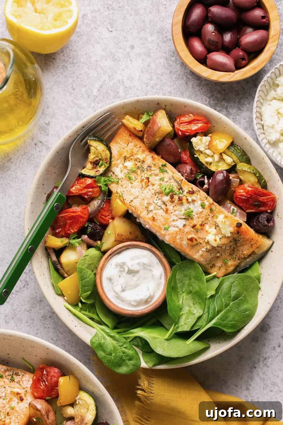 One Pan Greek Salmon with Roasted Vegetables 2 a bowl filled with veggies and spinach topped with a piece of greek salmon and tzatziki sauce.