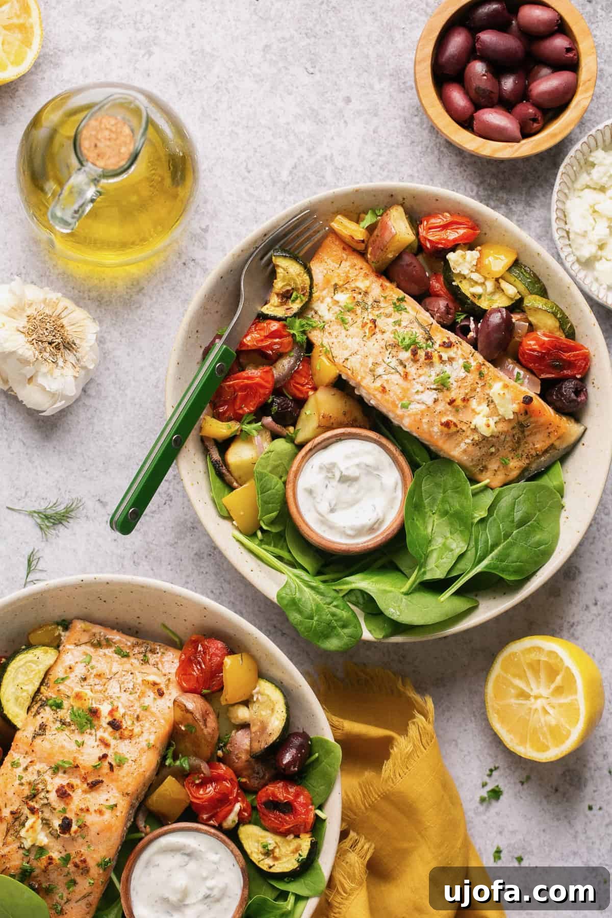 One Pan Greek Salmon with Roasted Vegetables 7 an overhead shot of two bowls of greek salmon with veggies and spinach and two small bowls of tzatziki sauce.