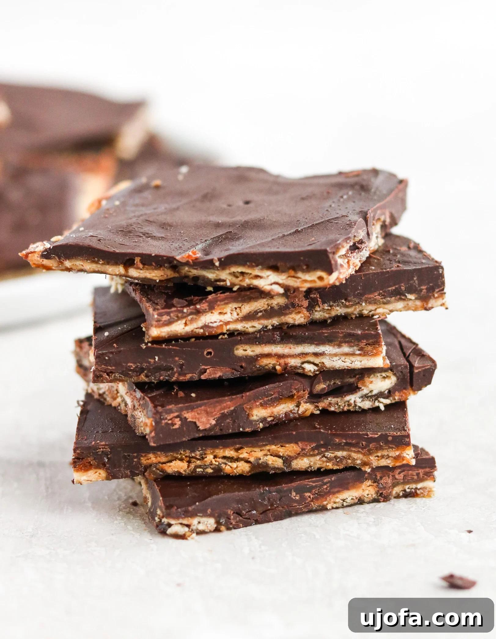 A stack of golden brown Christmas Crack dessert bars, featuring a crunchy cracker base, a glistening caramel layer, and a smooth dark chocolate topping, with festive holiday decorations in the background.