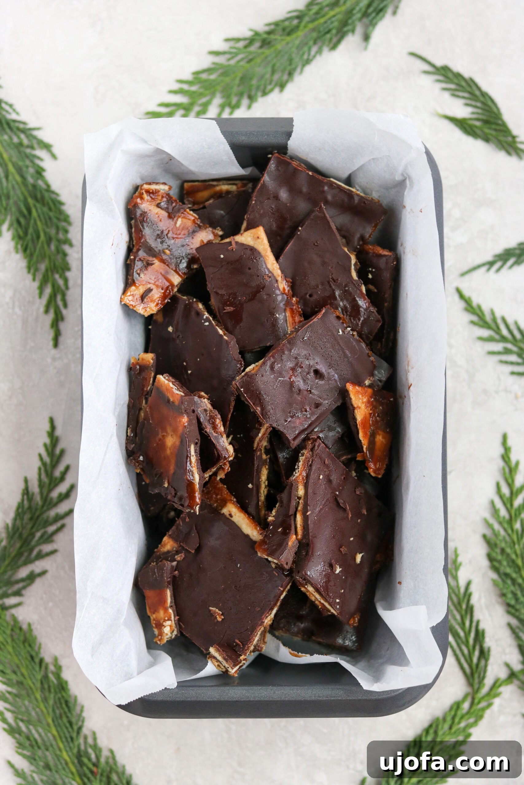 A festive loaf pan lined with parchment paper, filled with glistening Paleo Christmas crack candy, adorned with pine leaves and small red berries, evoking a cozy holiday atmosphere.