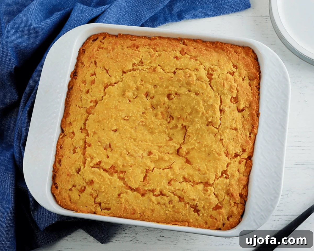 Golden baked gluten-free corn casserole in a white square baking dish, resting on a blue cloth with a lightly cracked, tender top.