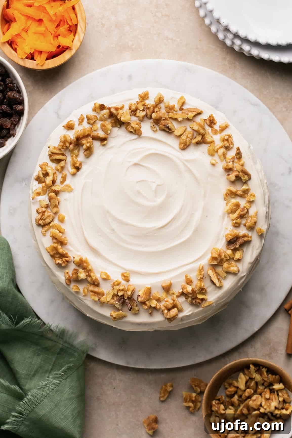 Decadent Gluten-Free Carrot Walnut Cake 8 An overhead shot of a beautifully frosted gluten-free carrot cake, adorned with walnuts, surrounded by fresh carrots, extra walnuts, a napkin, and plates, ready to be served.