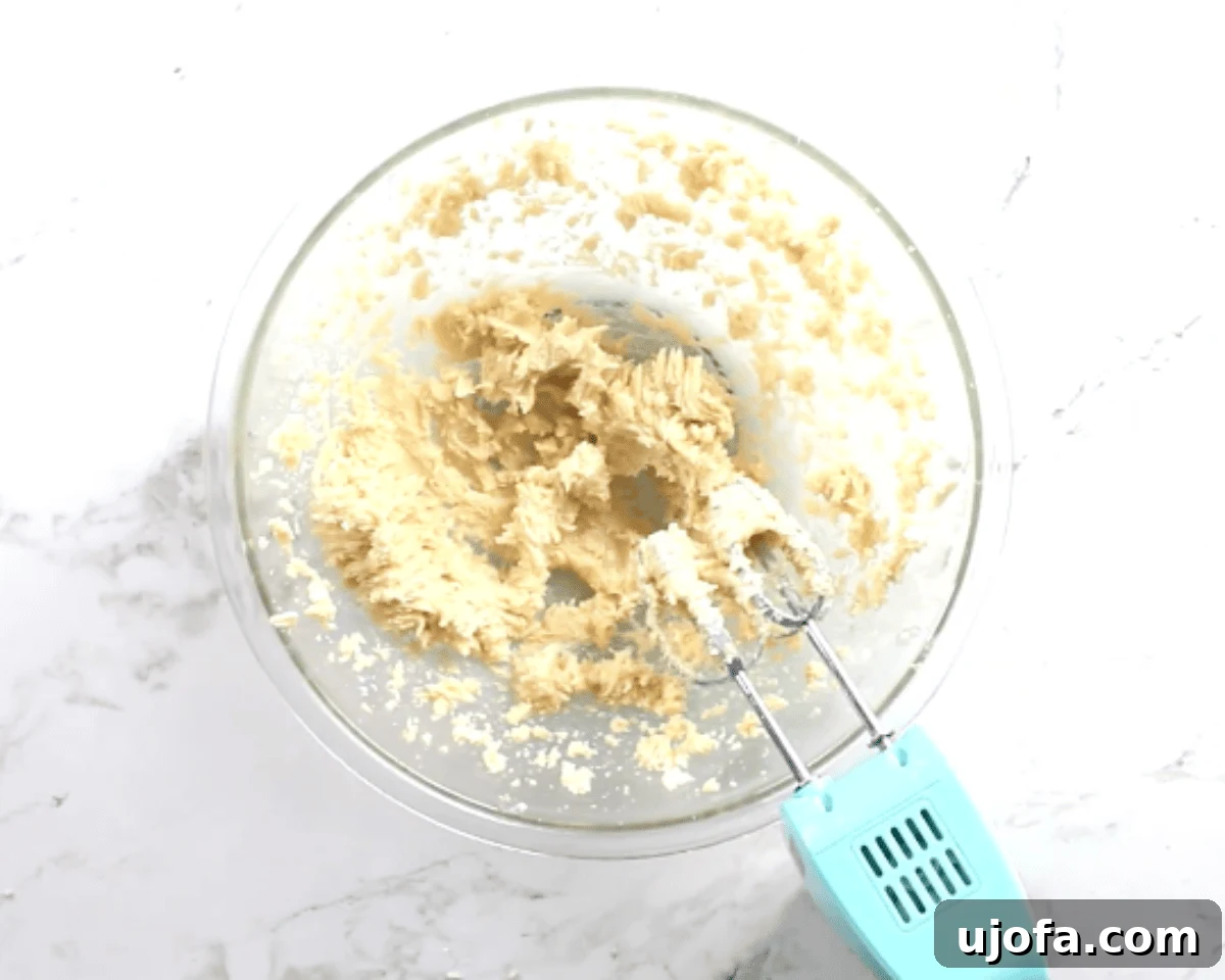 Creamed butter and sugars in a glass mixing bowl with an electric hand mixer on a marble countertop.