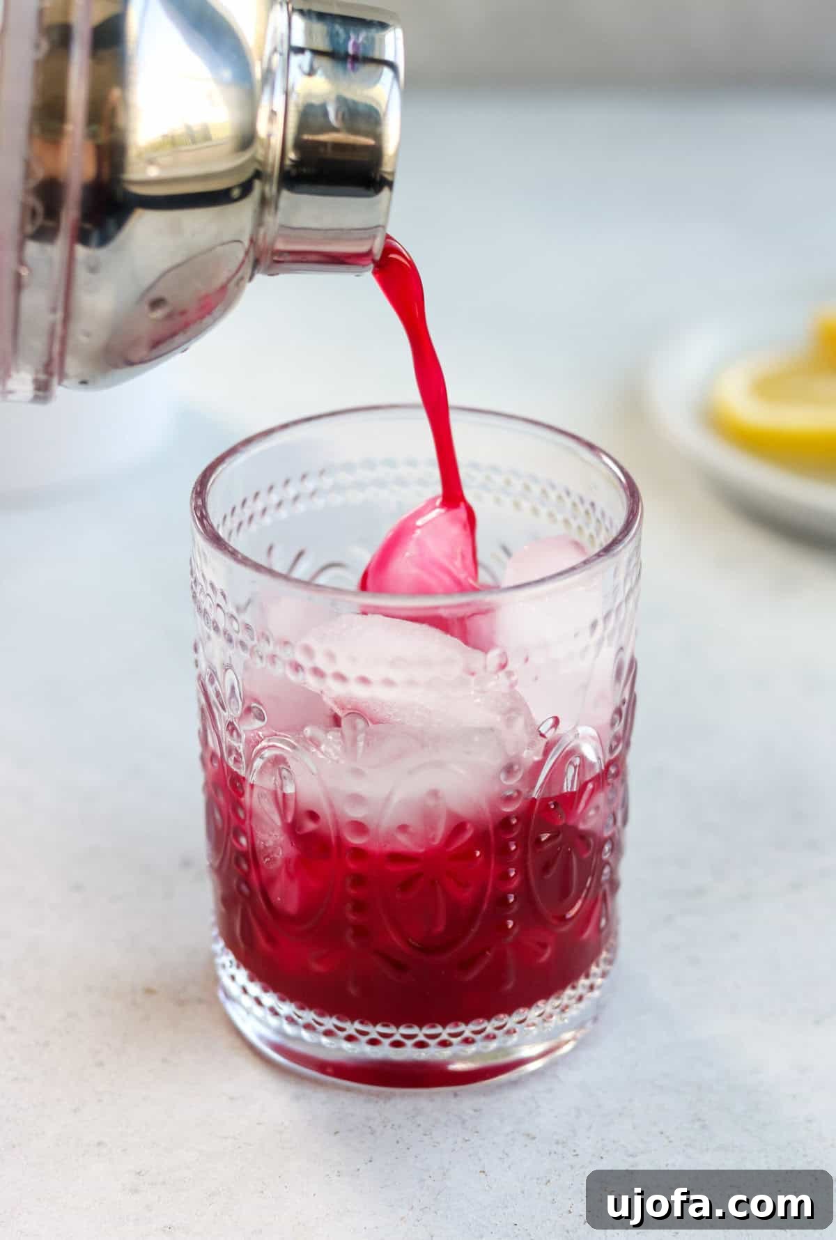 A cocktail shaker straining a vibrant lemon blueberry mocktail into a clear glass filled with ice cubes, capturing the moment of preparation.