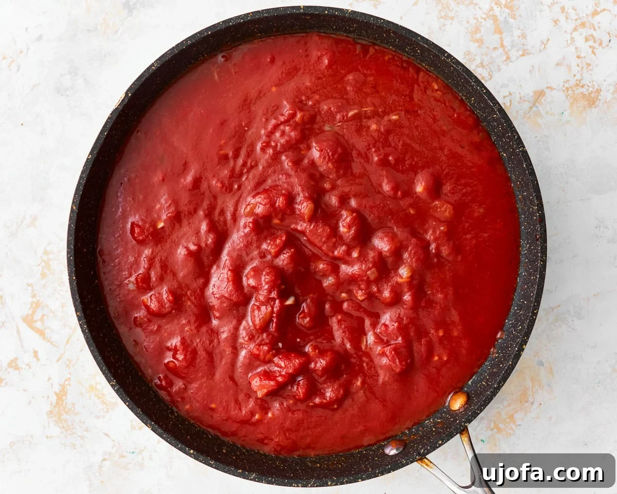 Crushed tomatoes and rich tomato sauce being poured into a skillet with sautéed aromatics, ready to be combined.