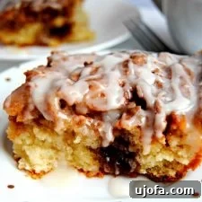 slice of gluten-free cinnamon roll cake on a white plate with another slice and coffee cup in the background