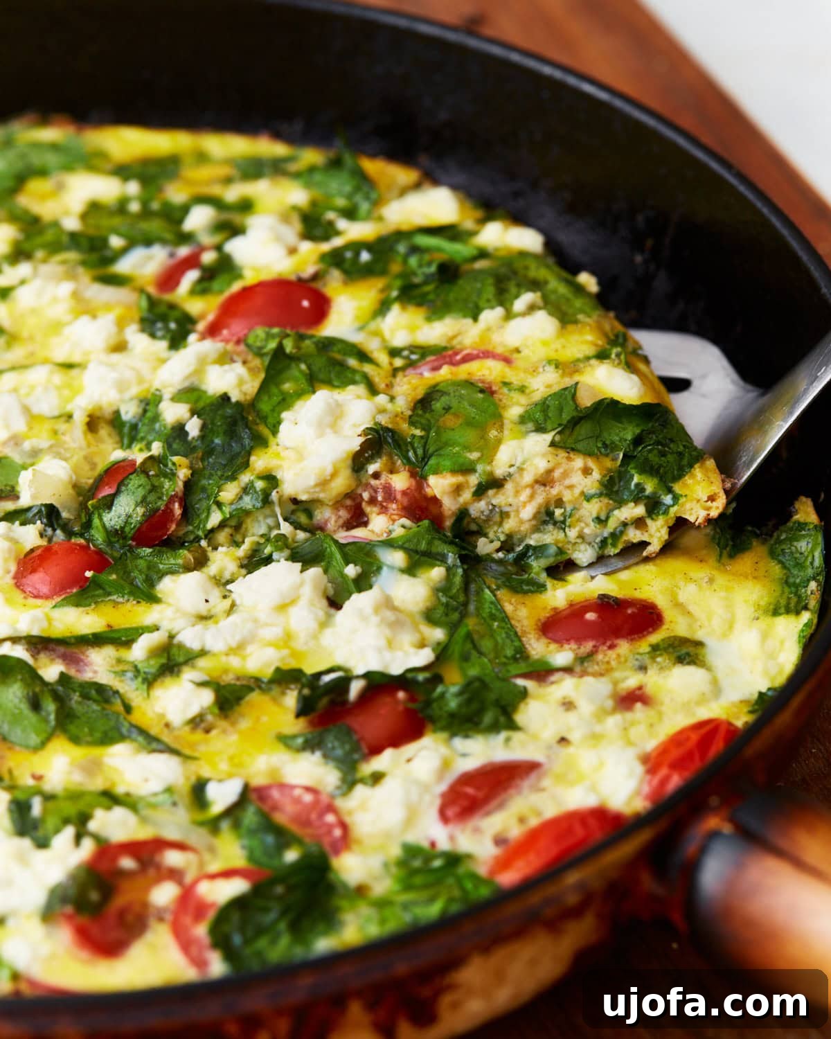 Savory Italian Egg Bake 2 A vibrant slice of gluten-free frittata with tomatoes, spinach, and feta cheese being lifted out of a black skillet by a metal spatula, showcasing its fluffy texture and colorful ingredients.