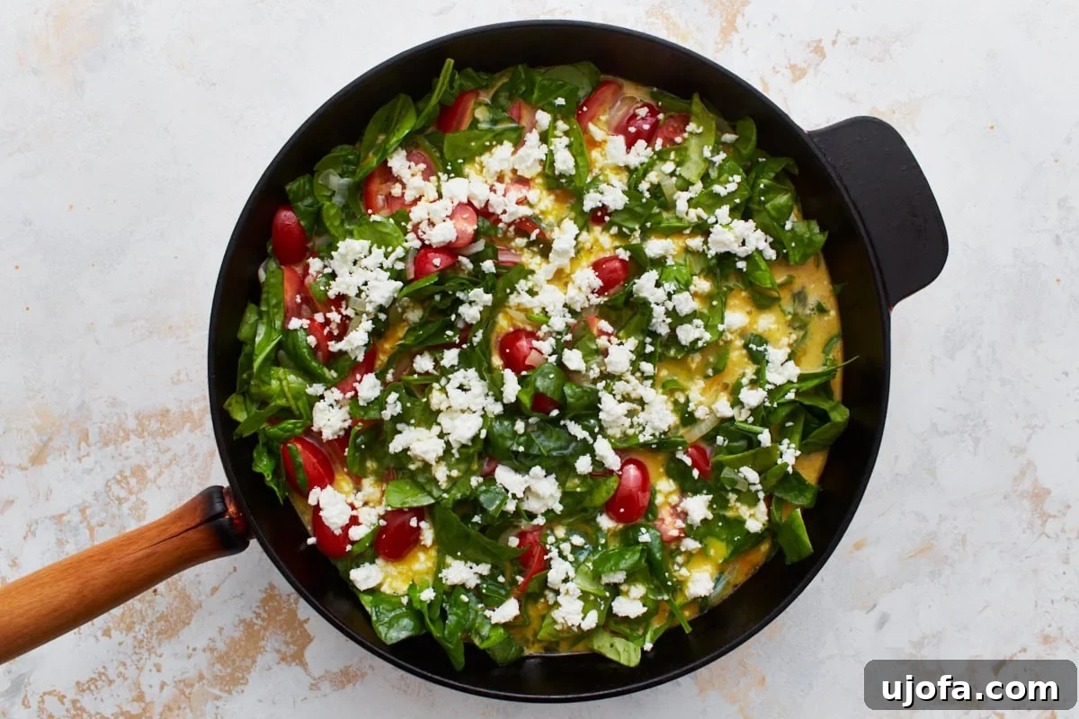Savory Italian Egg Bake 10 A vibrant, uncooked frittata mixture generously topped with fresh spinach, succulent cherry tomatoes, and crumbled feta cheese, beautifully arranged in a classic cast iron skillet.
