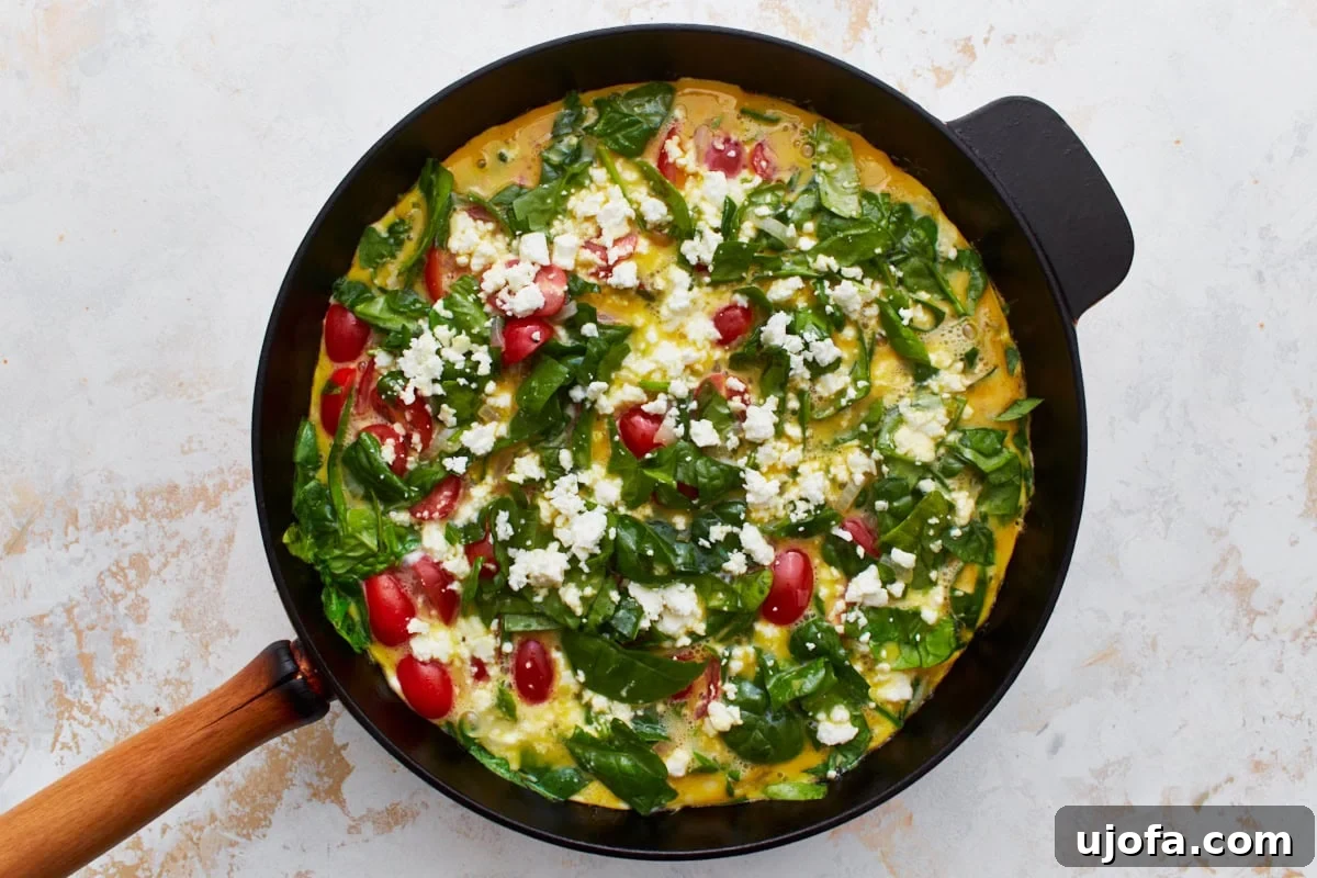 Savory Italian Egg Bake 11 A cast iron skillet holds a frittata that is just beginning to cook, with the edges of the egg mixture setting around the fresh spinach, cherry tomatoes, and crumbled feta cheese on top.