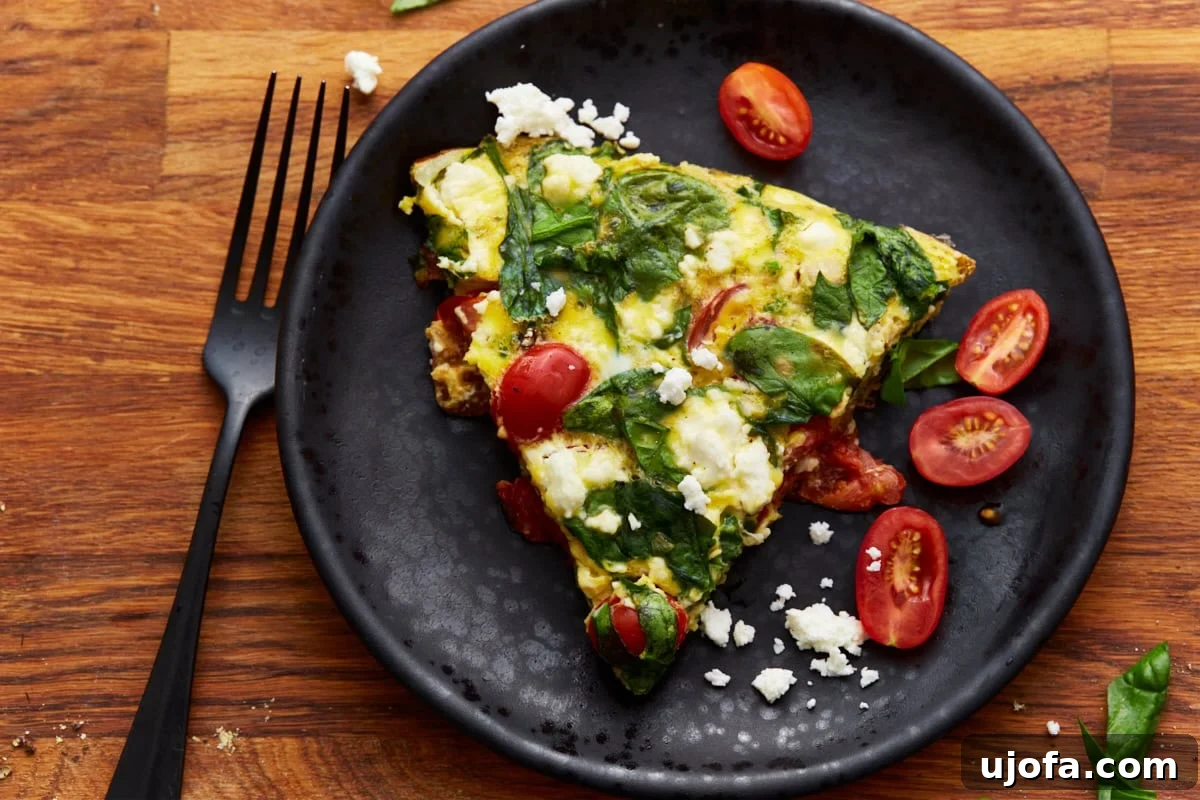 Savory Italian Egg Bake 14 A delicious slice of baked frittata, adorned with fresh spinach, juicy tomatoes, and savory feta cheese, elegantly presented on a black plate with a matching black fork. The meal is set on a rich wooden surface, enhancing its rustic appeal.