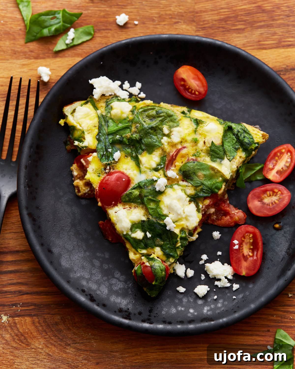Savory Italian Egg Bake 15 A delectable slice of baked frittata, brimming with spinach, tomatoes, and feta cheese, served on a black plate with a matching black spoon resting alongside, all set on a warm wooden surface.