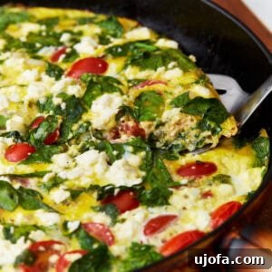 slice of frittata with tomatoes, spinach, and feta cheese being lifted out of black skillet by a metal spatula