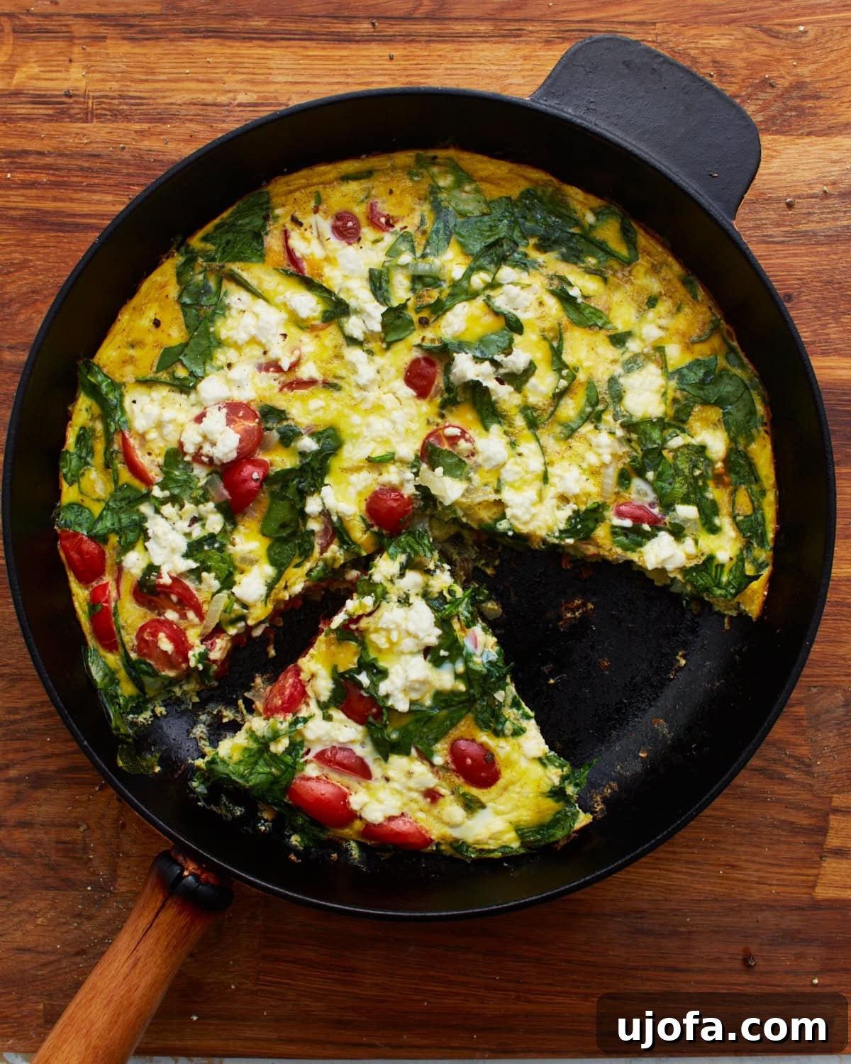Savory Italian Egg Bake 4 A beautifully baked frittata adorned with fresh spinach, halved grape tomatoes, and crumbled feta cheese, residing in a cast-iron skillet. A single slice has been carefully removed, showcasing the delightful layers and textures within. The skillet is artfully placed on a rustic wooden surface.