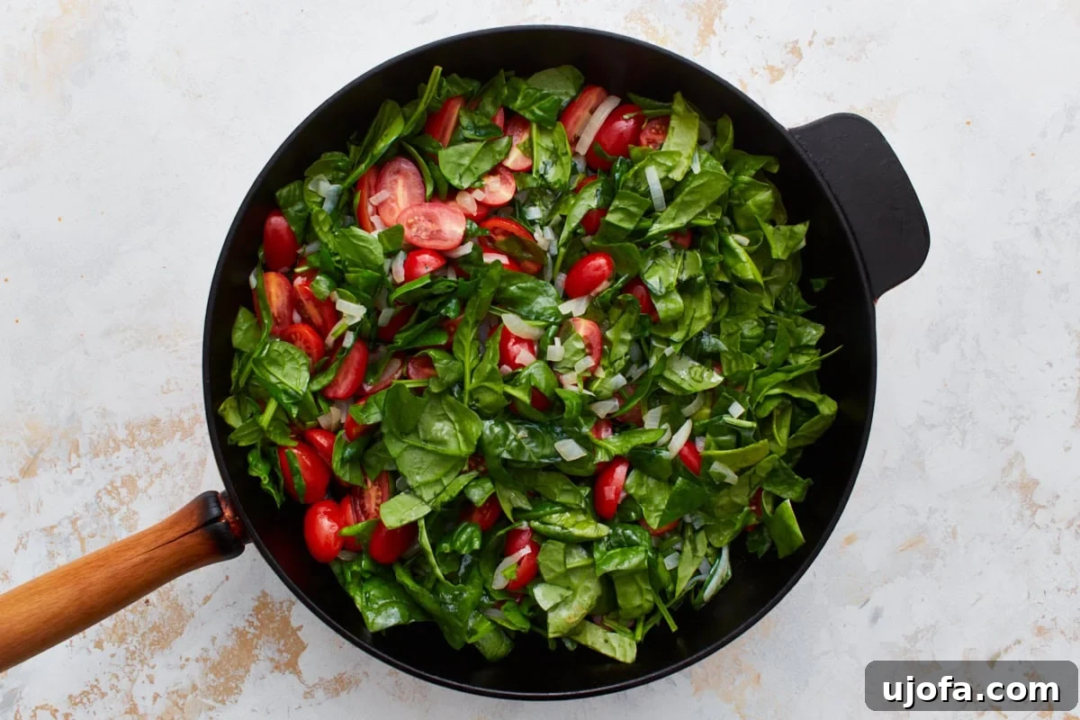 Savory Italian Egg Bake 9 A harmonious blend of fresh, vibrant chopped spinach and juicy halved grape tomatoes arranged invitingly in a black skillet with a sturdy wooden handle, set against a pristine light background.