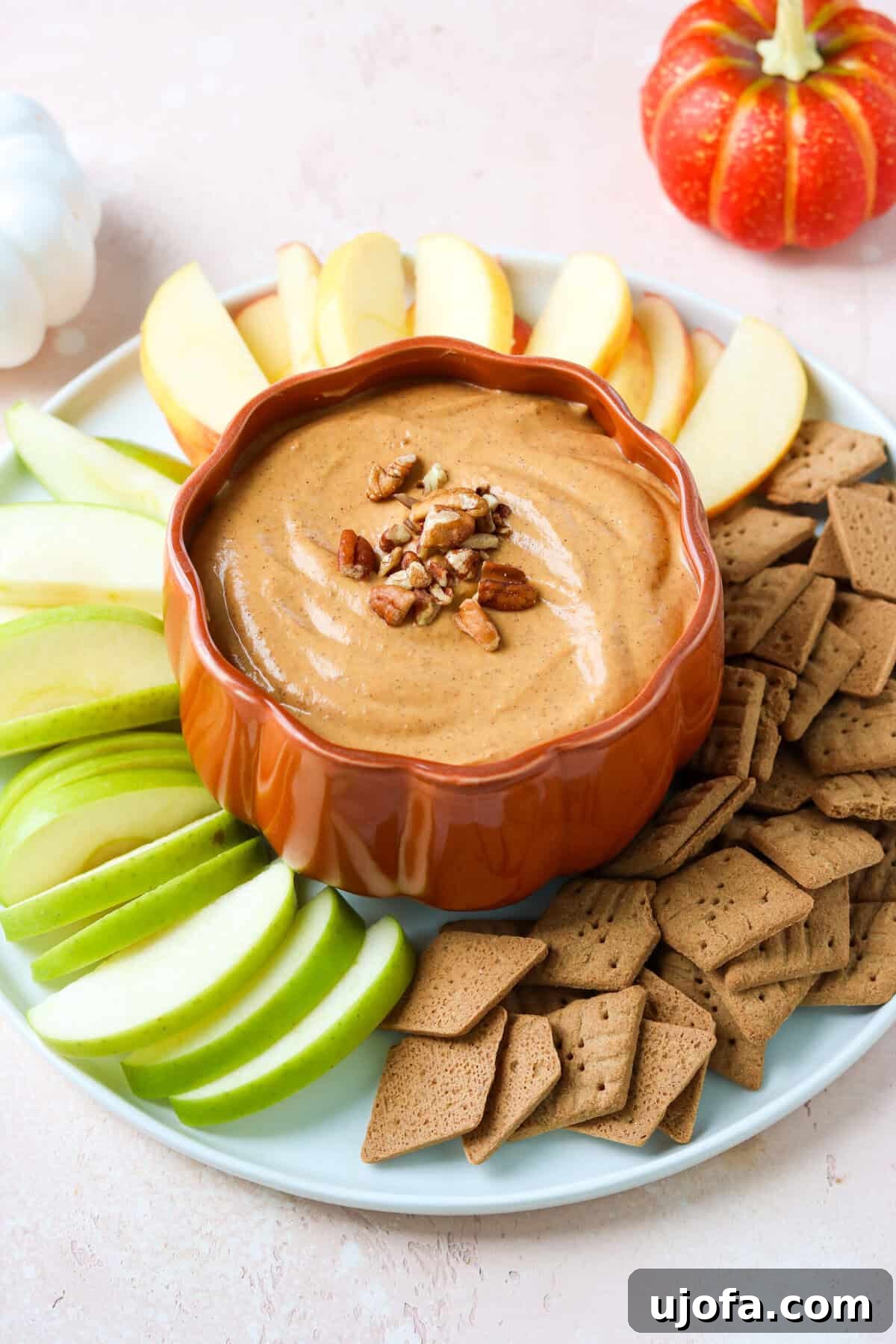 A vibrant pumpkin cream cheese dip served in a charming pumpkin-shaped bowl, surrounded by fresh apple slices and gluten-free sweet thins, evoking a cozy autumn atmosphere.