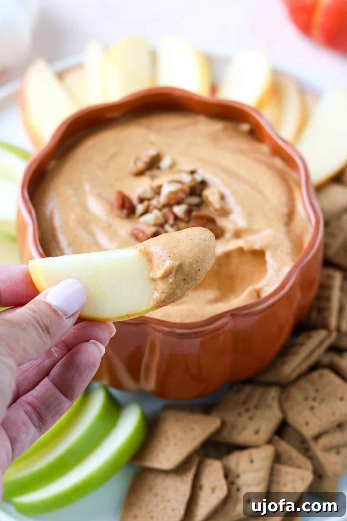 A hand gently dips a crisp apple slice into the creamy, orange pumpkin pie dip, highlighting the dip's appealing texture and inviting presentation.