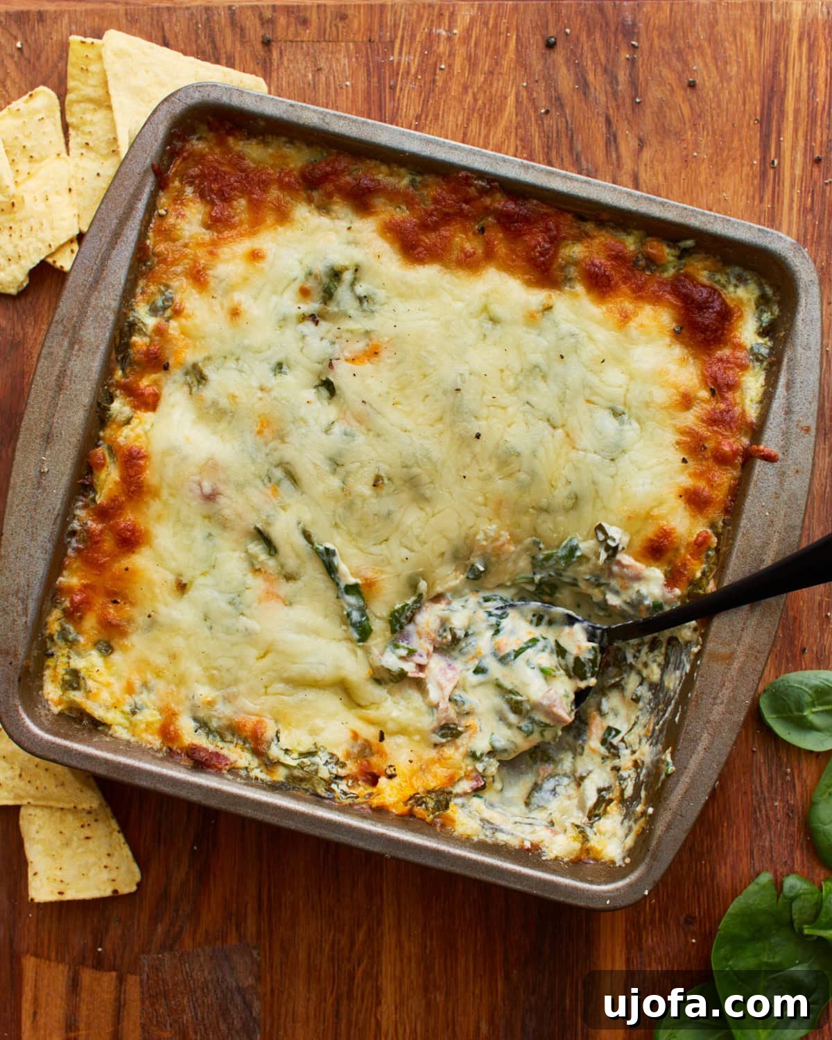 Velvety Warm Spinach Indulgence 2 overhead shot of a spoon dipped into a tin square baking dish with hot spinach dip topped with mozzarella cheese on a wooden surface