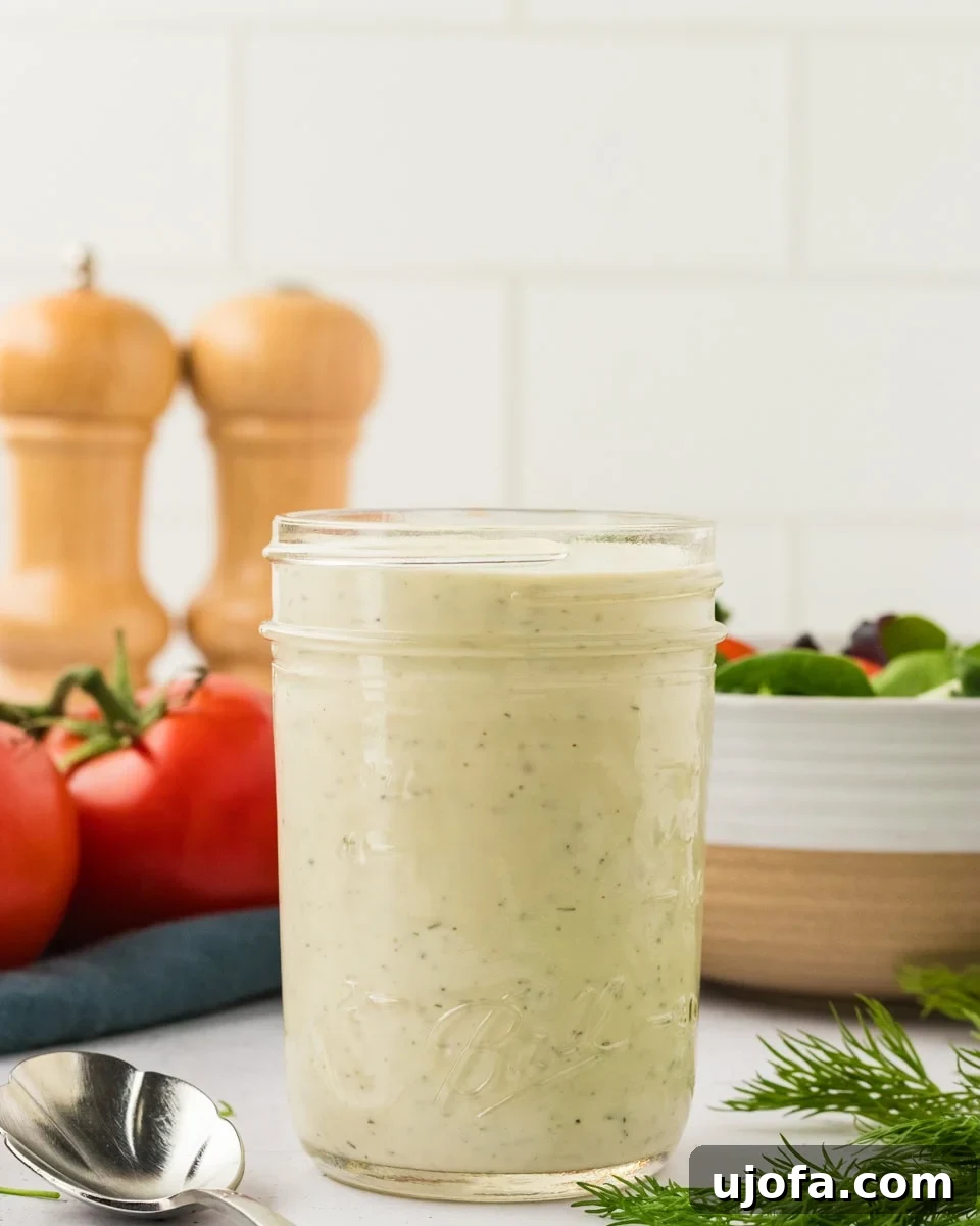 Luscious Plant-Based Ranch 2 A glass mason jar filled with creamy dairy-free ranch dressing, set on a white countertop with tomatoes, fresh dill, and a spoon nearby.