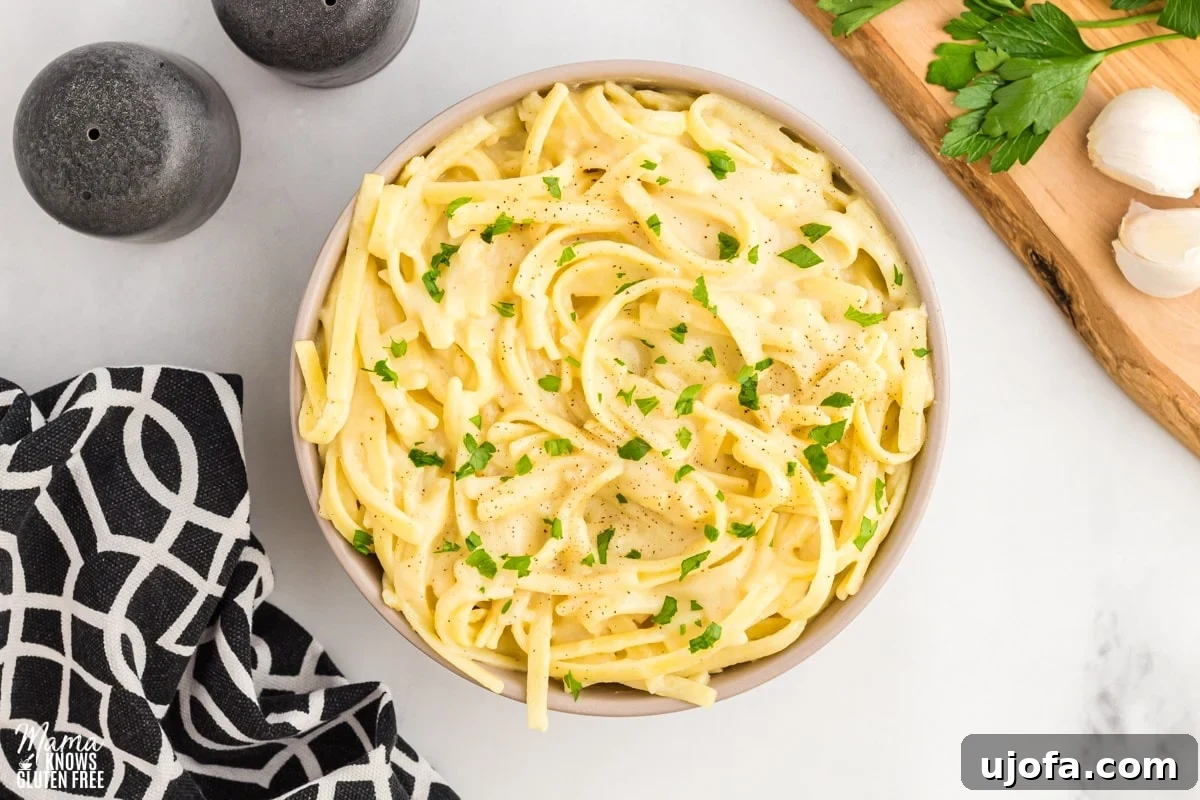 Velvety Plant-Based Alfredo 2 Creamy dairy-free Alfredo sauce coating pasta in an elegant cream bowl, surrounded by a black napkin, salt and pepper shakers, and a rustic cutting board.