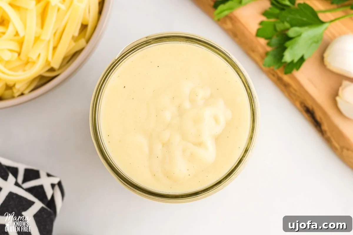 Velvety Plant-Based Alfredo 5 Dairy-free Alfredo sauce stored in a glass jar, with pasta, a black napkin, and a cutting board in the background, showcasing its rich texture.