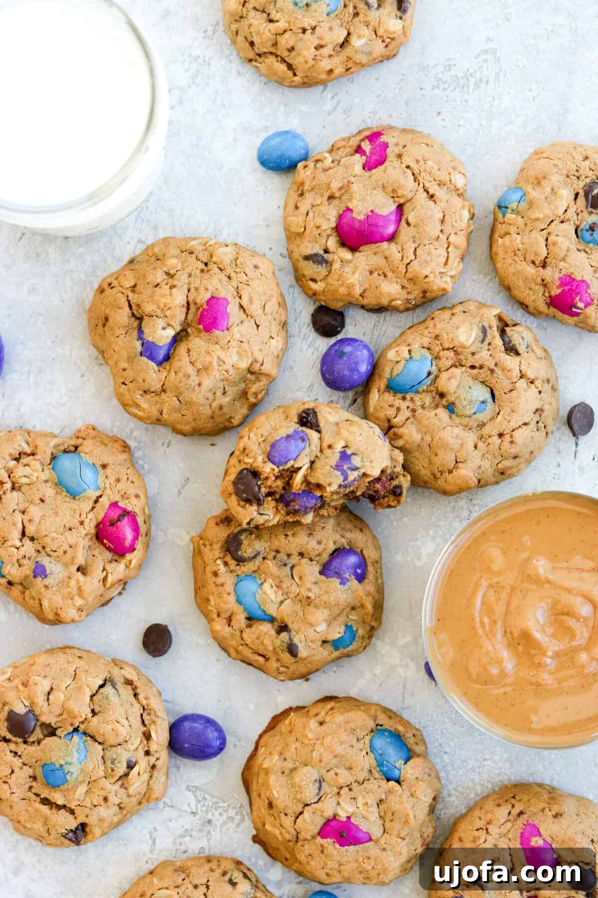 Gluten-free monster cookies with colorful chocolate candies, a glass of milk, and a bowl of peanut butter, highlighting their delicious and appealing nature.