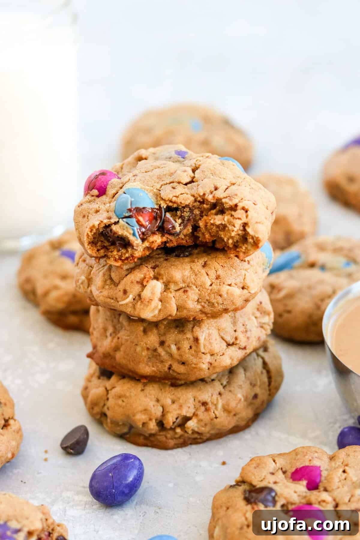 A stack of four gluten-free monster cookies surrounded by colorful M&Ms and other cookies, showcasing their rich texture and appealing mix-ins.