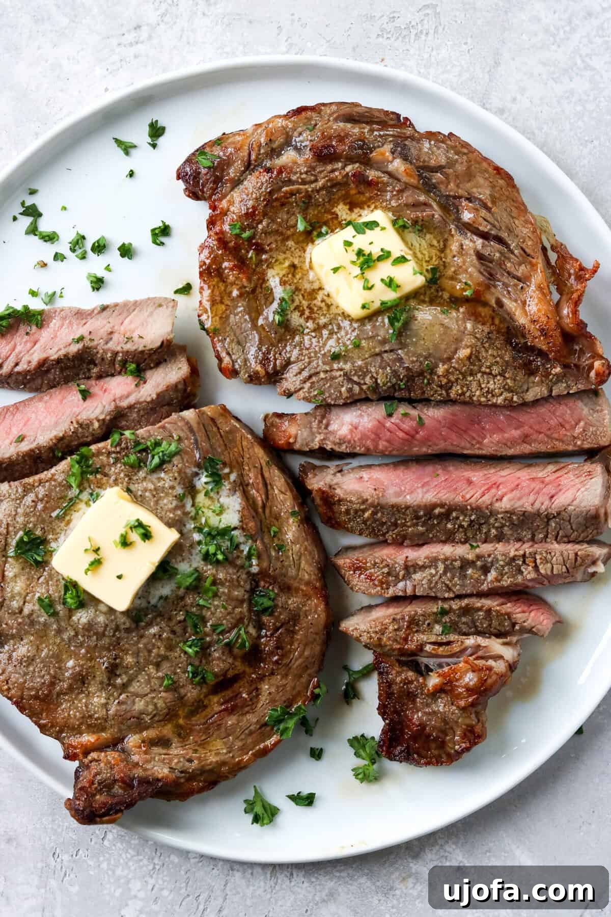 Sliced ribeye steak on a white plate, artfully topped with a melting pat of butter and fresh parsley, ready to be savored.