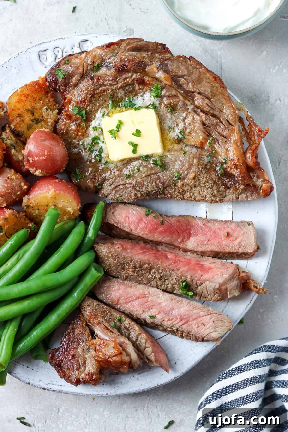 Succulent sliced ribeye steak, richly topped with butter and parsley, presented with vibrant green beans and golden roasted potatoes on the side.