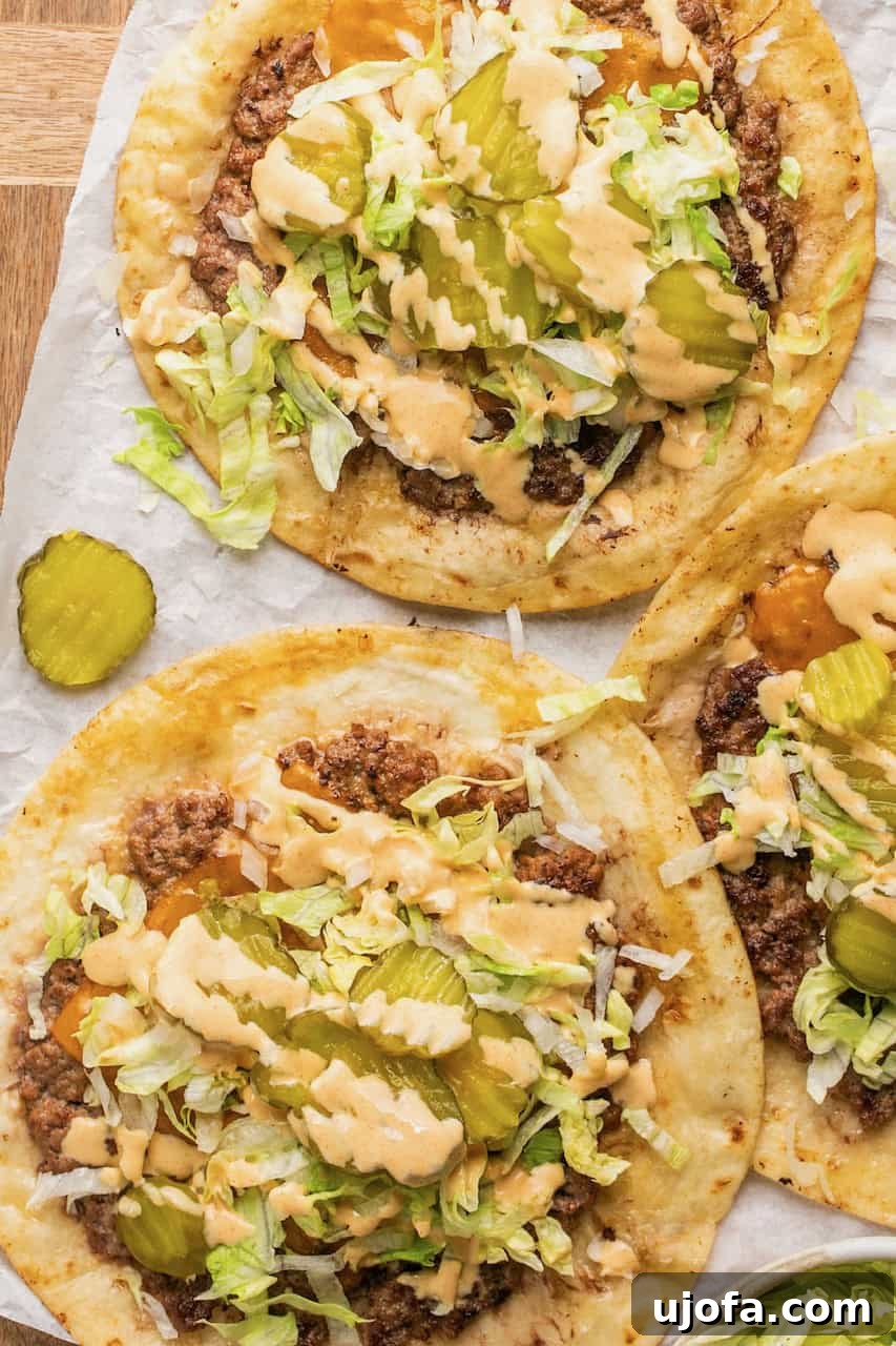Close-up of three smash burger Big Mac tacos on parchment paper, beautifully garnished with shredded lettuce, sliced pickles, melted cheese, and a swirl of big mac sauce.