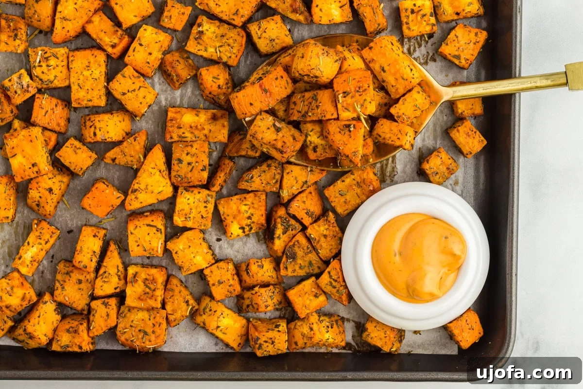 Golden Roasted Sweet Potatoes 9 Roasted sweet potato cubes on a baking sheet with a golden spoon and a small white bowl of creamy dipping sauce.