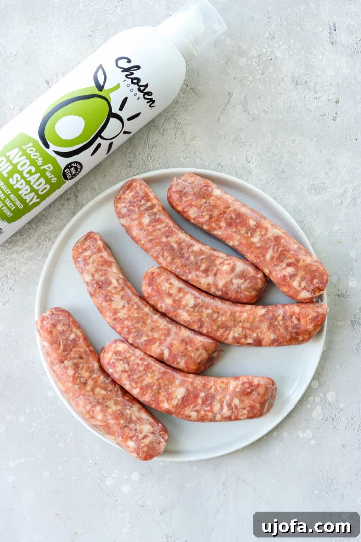 Quick and Crispy Air Fryer Italian Sausage 3 Raw Italian sausages on a white plate, with a bottle of avocado oil spray nearby, ready for cooking.
